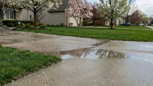 An aging concrete driveway in a suburban neighborhood with standing water puddles and poor drainage during a Michigan spring.