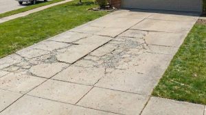 A residential concrete driveway with extensive cracks and winter damage, with a yard sign asking "Repair or Replace?" in a spring suburban neighborhood.